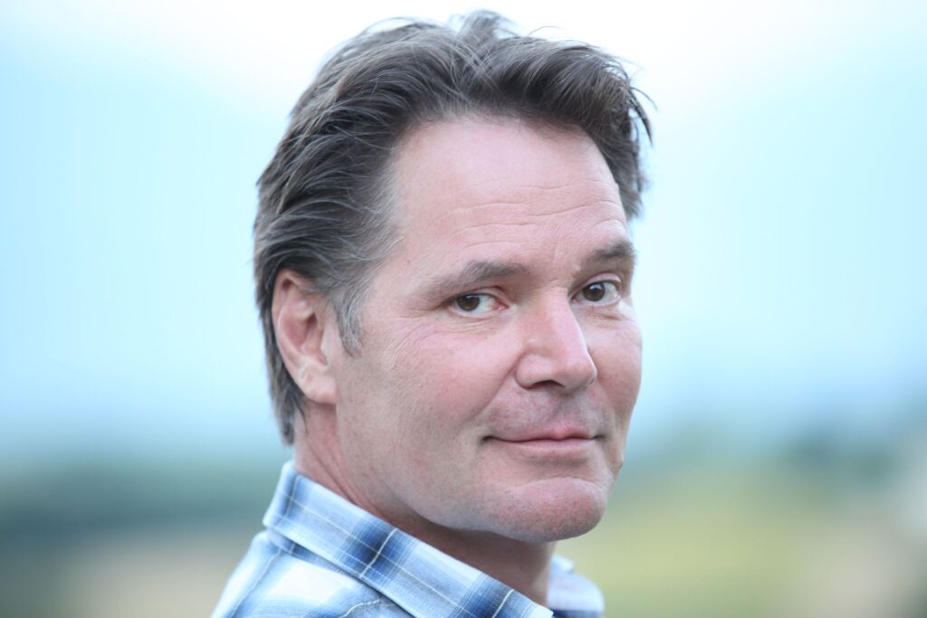 David Parsons looks over his right shoulder directly at the camera, the headshot nearly filling the frame. He has White skin and is clean-shaven, with a small smile at the corners of his mouth and his left eyebrow raised slightly. His hair is short, brown, and swept over his forehead and behind his visible right ear. The collar of his blue and white plaid shirt stands out against the pale greens and blues of the blurred outdoor landscape behind him.