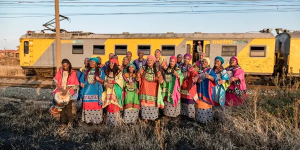 Soweto Gospel Choir members in colorful attire, singing outdoors with a yellow train behind them.