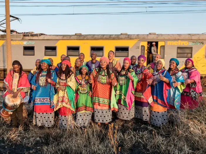 Soweto Gospel Choir members in colorful attire, singing outdoors with a yellow train behind them.