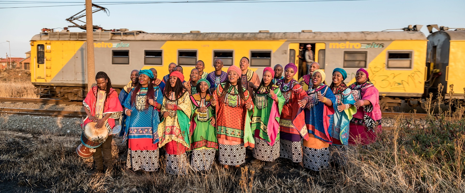 Soweto Gospel Choir members in colorful attire, singing outdoors with a yellow train behind them.