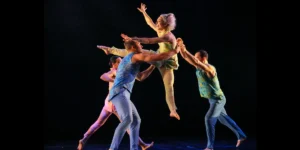 Four Pilobolus dancers pictured on stage, including one dancer smiling while jumping in the air in middle of frame.