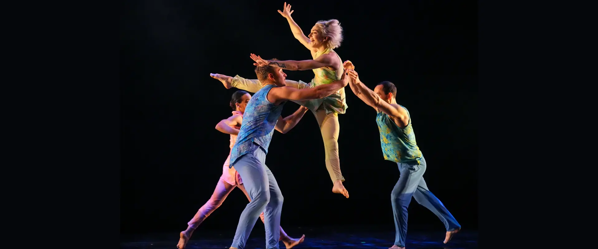 Four Pilobolus dancers pictured on stage, including one dancer smiling while jumping in the air in middle of frame.