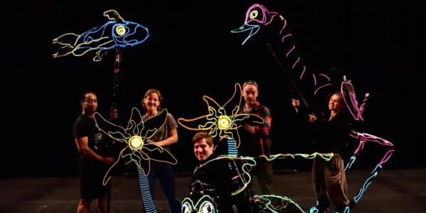 A group of smiling puppeteers with their colorful, glowing animal creations against a dark background.