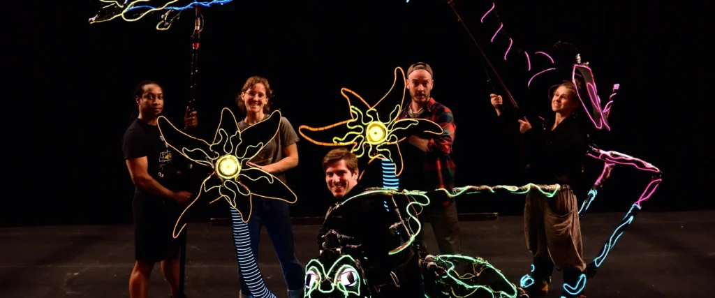 A group of smiling puppeteers with their colorful, glowing animal creations against a dark background.