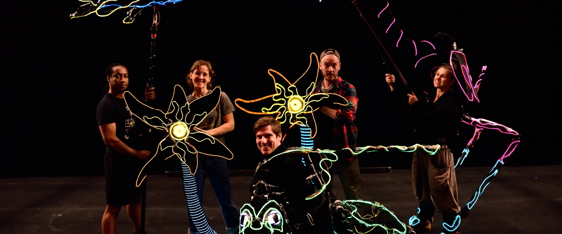 A group of smiling puppeteers with their colorful, glowing animal creations against a dark background.