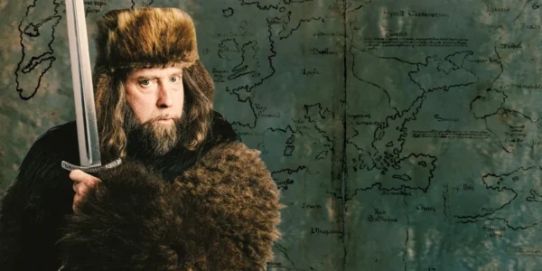 Comedian Josh Blue in a fur hat and coat, holding a sword, in front of an old map.