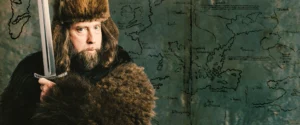 Comedian Josh Blue in a fur hat and coat, holding a sword, in front of an old map.