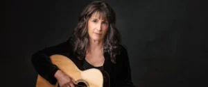 Singer-songwriter Karla Bonoff with her acoustic guitar, looking directly at the camera. For her concert.