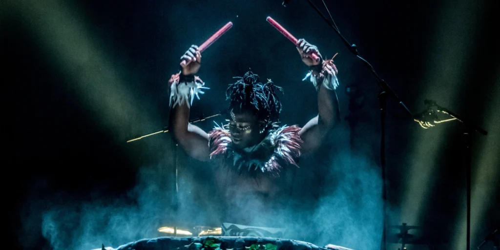 A performer with painted face and feathers, raising drumsticks into bright stage lights. For Cirque Kalabanté.
