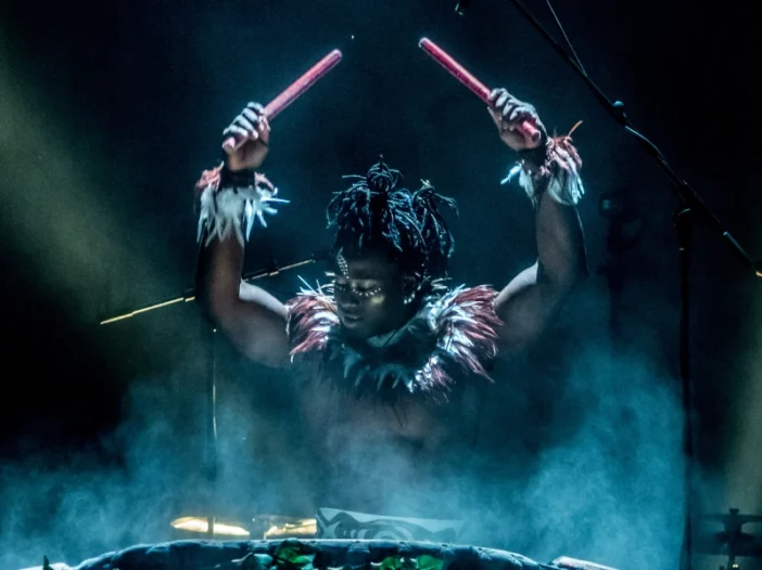 A performer with painted face and feathers, raising drumsticks into bright stage lights. For Cirque Kalabanté.