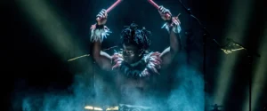 A performer with painted face and feathers, raising drumsticks into bright stage lights. For Cirque Kalabanté.