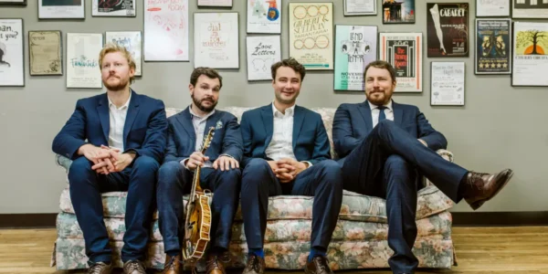 Four musicians, members of The Slocan Ramblers, in blue suits, casually seated on a floral couch in front of a wall of framed artwork.