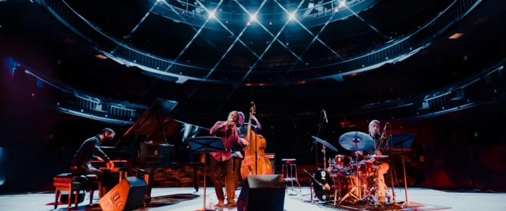 LP & The Vinyl performing jazz on stage with piano, bass, and drums under spotlights. For their concert.