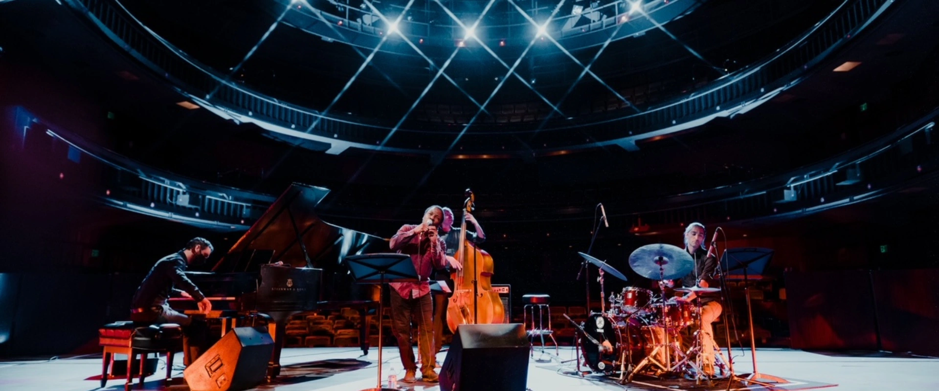 LP & The Vinyl performing jazz on stage with piano, bass, and drums under spotlights. For their concert.