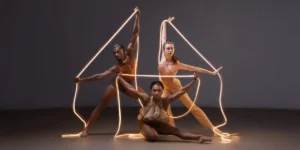 Three dancers from the Ailey II dance company, posed with illuminated string lighting as props.