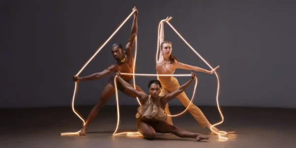 Three dancers from the Ailey II dance company, posed with illuminated string lighting as props.