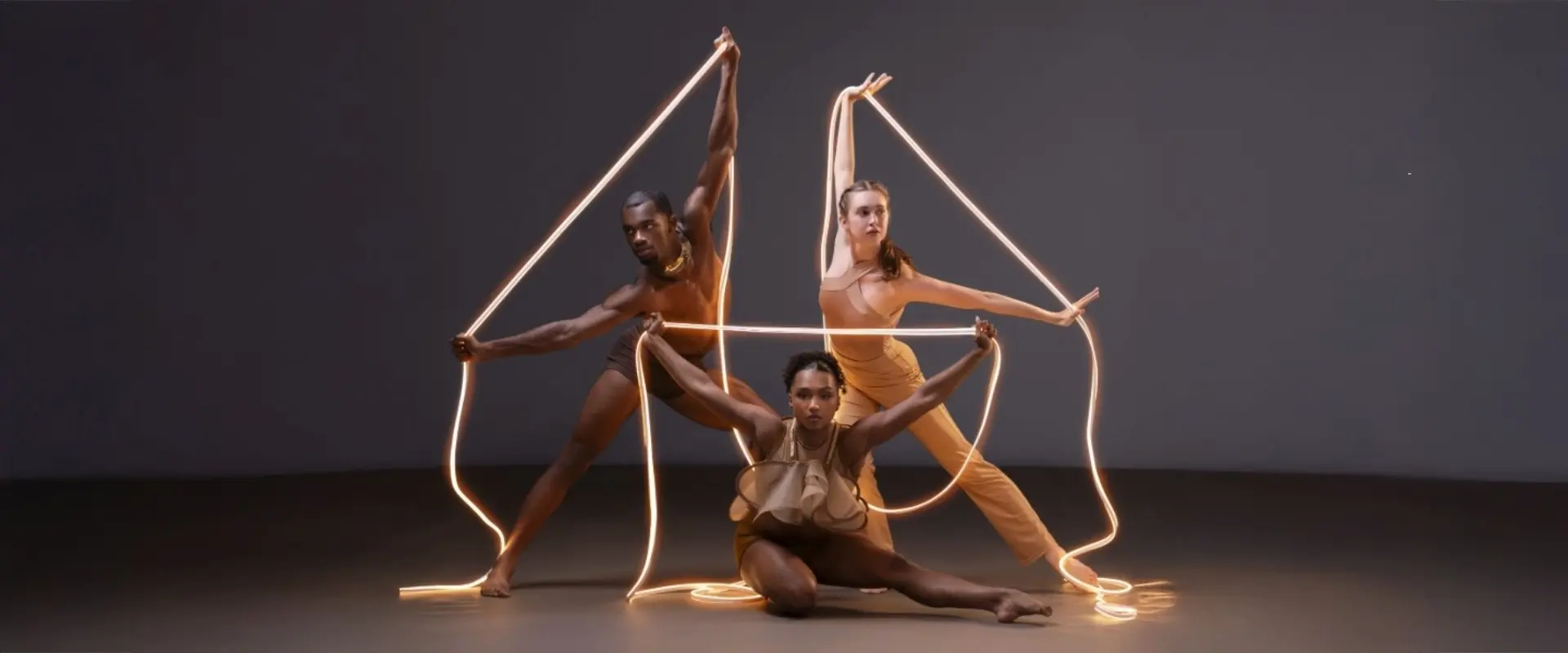 Three dancers from the Ailey II dance company, posed with illuminated string lighting as props.