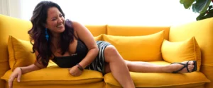 Jazz singer-songwriter Nicole Zuraitis in a striped skirt and black top, relaxing and smiling on a yellow couch.