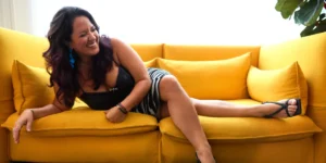 Jazz singer-songwriter Nicole Zuraitis in a striped skirt and black top, relaxing and smiling on a yellow couch.