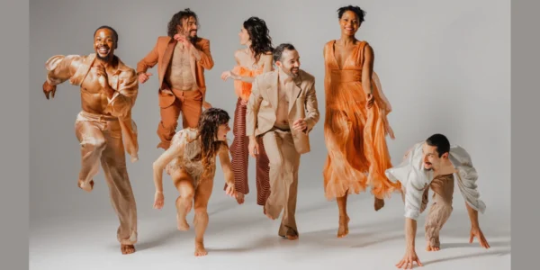 Six BODYTRAFFIC dancers in various contemporary dance movements, some mid-air, in earth-toned outfits.