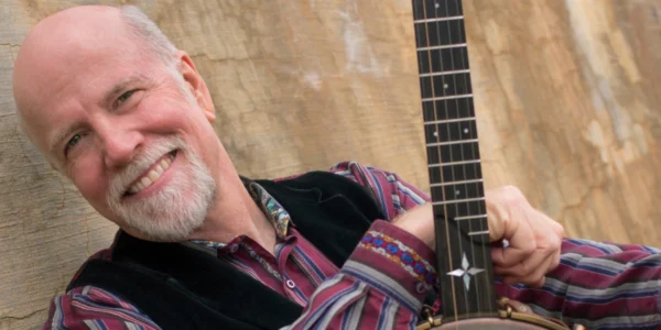 John McCutcheon, folk artist, leaning with his banjo against a textured wall. Image for his performance.