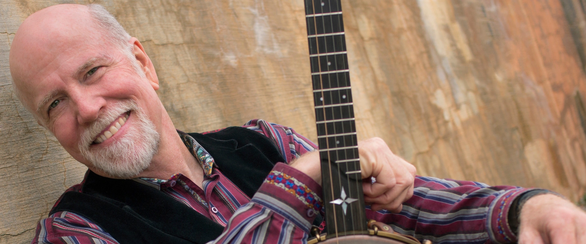 John McCutcheon, folk artist, leaning with his banjo against a textured wall. Image for his performance.