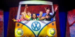 Cast of "Pete the Cat," including a character with cat ears, making peace signs from a yellow and blue VW bus prop.