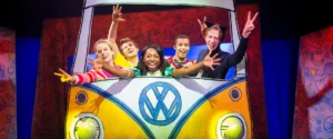 Cast of "Pete the Cat," including a character with cat ears, making peace signs from a yellow and blue VW bus prop.