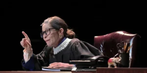 Actor portraying Ruth Bader Ginsburg in judicial robes, seated at a desk, pointing. For "All Things Equal" play.