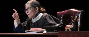 Actor portraying Ruth Bader Ginsburg in judicial robes, seated at a desk, pointing. For "All Things Equal" play.