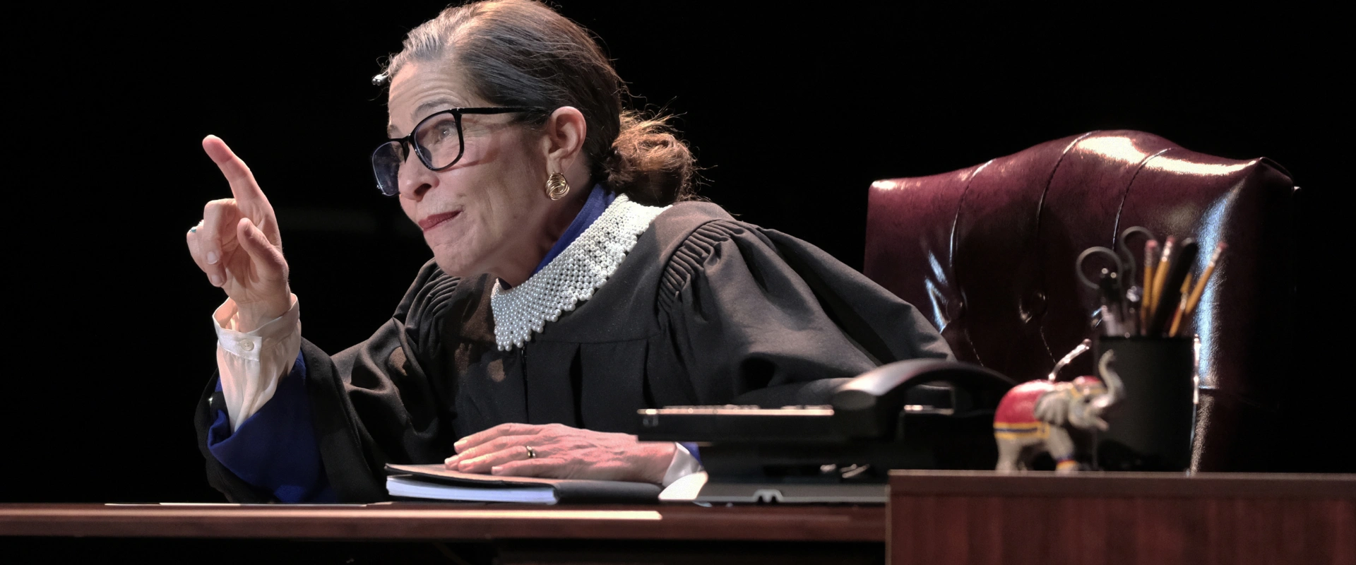 Actor portraying Ruth Bader Ginsburg in judicial robes, seated at a desk, pointing. For 
