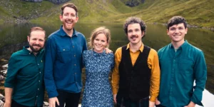 The five members of the band Breabach smiling outdoors, standing by a lake with mountains behind them.