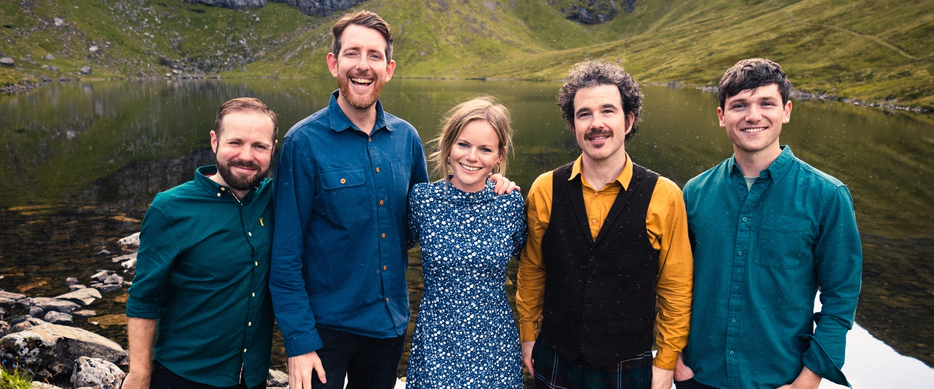 The five members of the band Breabach smiling outdoors, standing by a lake with mountains behind them.