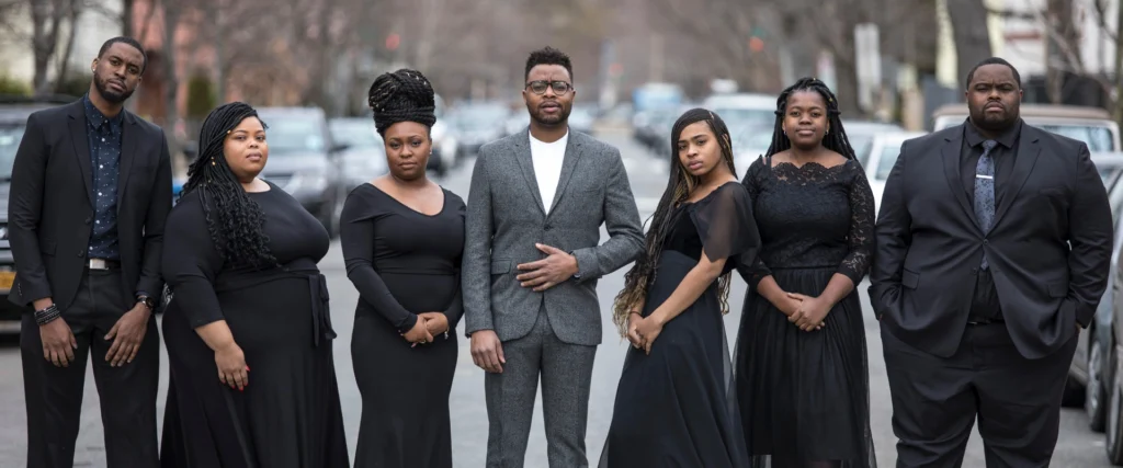Reginald Golden in a grey suit, flanked by six singers in black dresses and suits on a street. Context: 