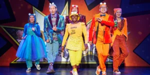 Cast of "Cat Kid Comic Club" in bright, mismatched outfits and playful poses, on stage with a comic book backdrop.