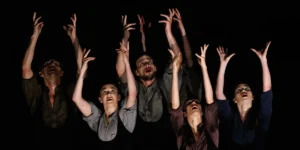 Eight dancers reaching upwards with expressive faces on a dark stage. On page for Stewart/Owen Dance + Guests.