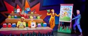Cast of "Cat Kid Comic Club" in bright, mismatched outfits and playful poses, on stage with a comic book backdrop.