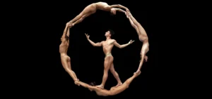 Photo: dancers posed linking arms in a circle with one dancer walking on the others inside the circle