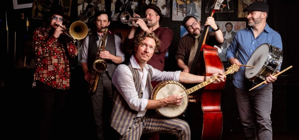 Blair Crimmins and his five piece band; Blair Crimmins in front with a banjo, a trombonist, saxophonist, trumpet player, upright bassist, and drummer