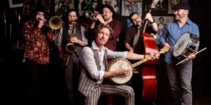 Blair Crimmins and his five piece band; Blair Crimmins in front with a banjo, a trombonist, saxophonist, trumpet player, upright bassist, and drummer