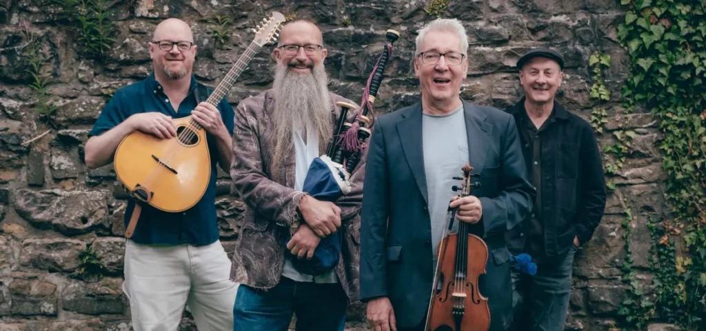 four band members of Old Blind Dogs smiling at camera; one holding a bazuki, one holding a fiddle, and one holding bagpipes