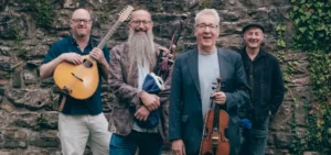 four band members of Old Blind Dogs smiling at camera; one holding a bazuki, one holding a fiddle, and one holding bagpipes